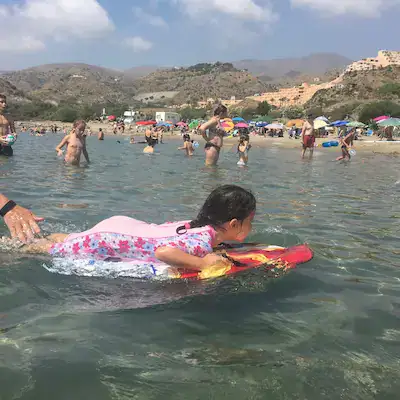 A child friendly beach in Mojácar in southern Spain, Showing a girl in a pink sun protection swimming costume, laying on a body board, with people in the background standing in the water. Hills in the background.