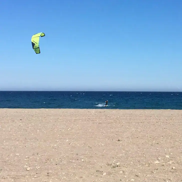 A beach in Mojácar with a kitesurfer with a green kite. The sky is blue, the sea is a deeper blue and the sand in the foreground has some small pebbles, but is mainly sand.