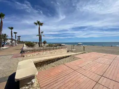 Accessible beach in Mojacar.  The paseo which is above sea level is shown on the left, has a concrete slope down to beach level that turns into a wooden "boardwalk" across the sand towards the shore.  There is also a fixed shelter providing shade.