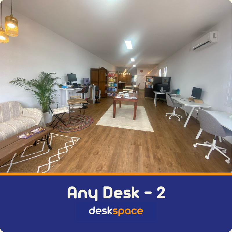 A picture taken from within DeskSpace Coworking Mojacar, Spain showing the view into the office. Wooden floor, natural coloured rugs on the floor and a sofa soften the square lines of the desks. The banner at the bottom says Any Desk - 2 and the DeskSpace logo.